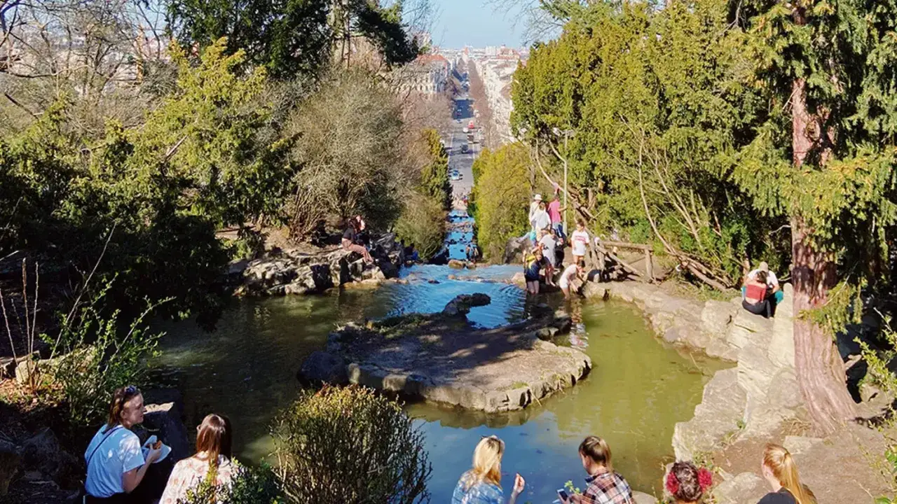 Menschen genie&szlig;en den sonnigen Tag am k&uuml;nstlichen wasserfall berlin, mit Blick auf eine belebte Stra&szlig;e im Hintergrund.