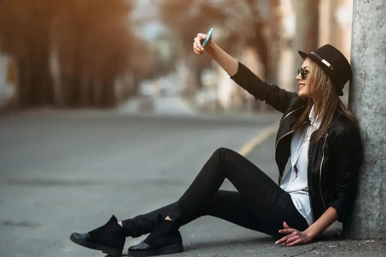 Kobieta w kapeluszu i skórzanej kurtce robi selfie telefonem, pokazując, jak nowoczesne środki masowego przekazu kształtują nasz świat.