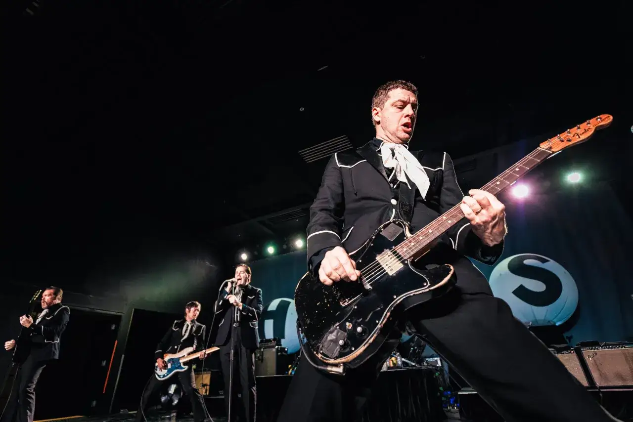 Miembros de The Hives enérgicos en el escenario, con trajes negros y corbatas blancas, tocando guitarras eléctricas.