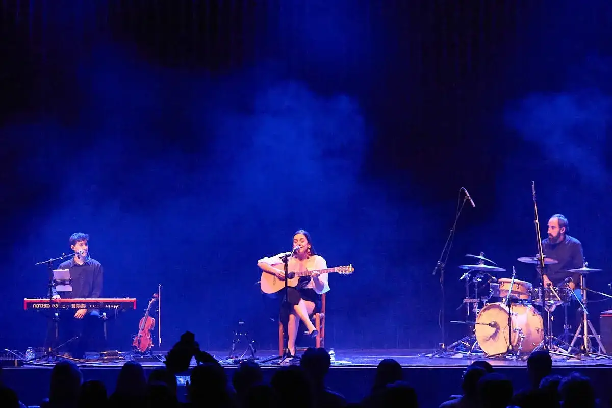 Valeria Castro Zaragoza canta y toca la guitarra en un escenario azul, acompañada por un teclista y un baterista.