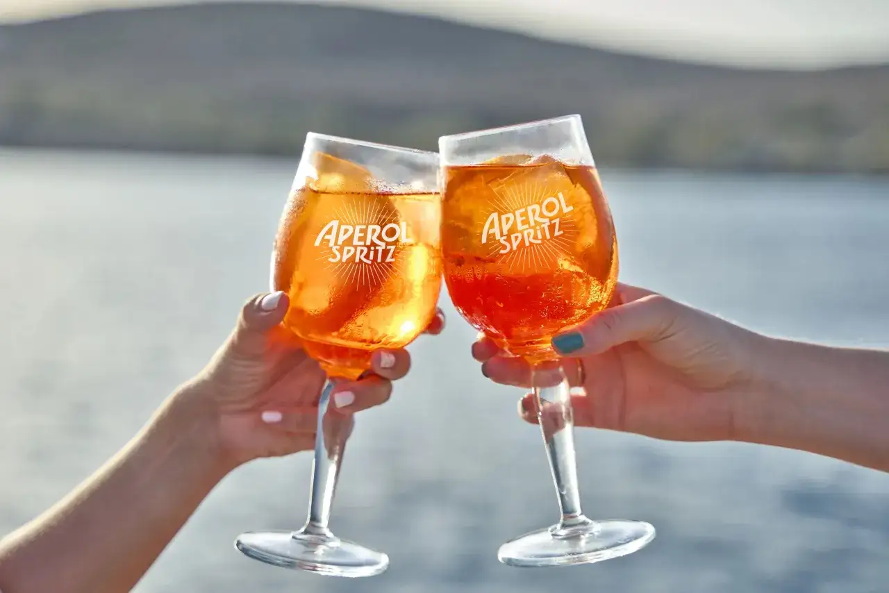 Zwei Hände stoßen mit Aperol Spritz Gläsern an. Das leuchtend orangefarbene Getränk mit Eis und Orangenscheibe ist perfekt für das Aperol Spritz Rezept.