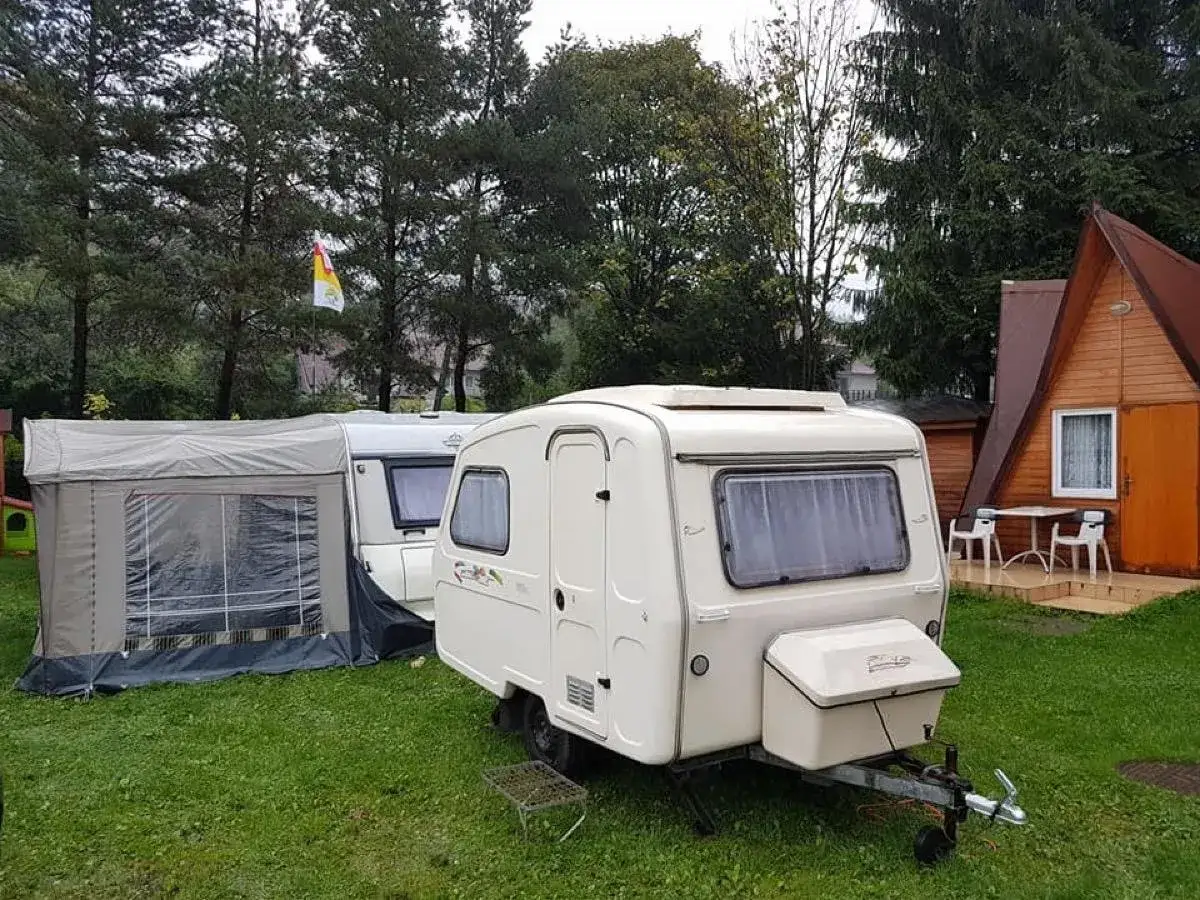 Dwa białe kampery i przyczepa kempingowa z przedsionkiem na zielonej trawie. W tle drewniany domek i drzewa. Idealne miejsce na camping Jonidło.