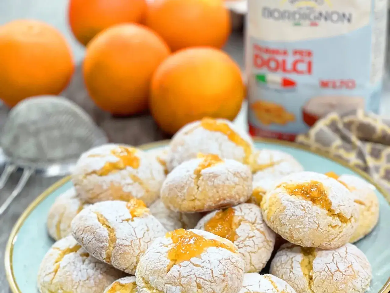 Biscotti morbidi all'arancia, cosparsi di zucchero a velo, con un ripieno dorato di marmellata. Sullo sfondo, arance fresche e una confezione di farina per dolci.