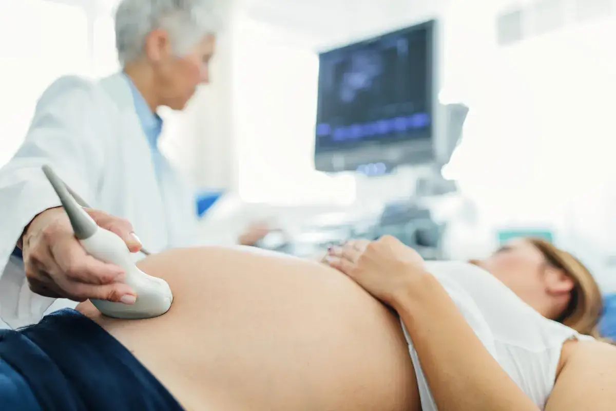 Ecografia di una donna incinta, con il medico che esegue l'esame. Si vede la pancia e il monitor con l'immagine del feto, probabilmente in posizione fetale a 27 settimane.