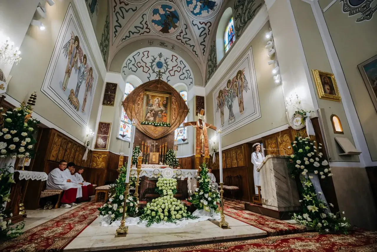 Wnętrze kościoła ozdobione kwiatami na uroczystość. Widać ołtarz, krzyż, malowidła na ścianach i suficie, a także ministrantów. W tle dziewczynka w białej sukience, być może na pierwszej komunii.