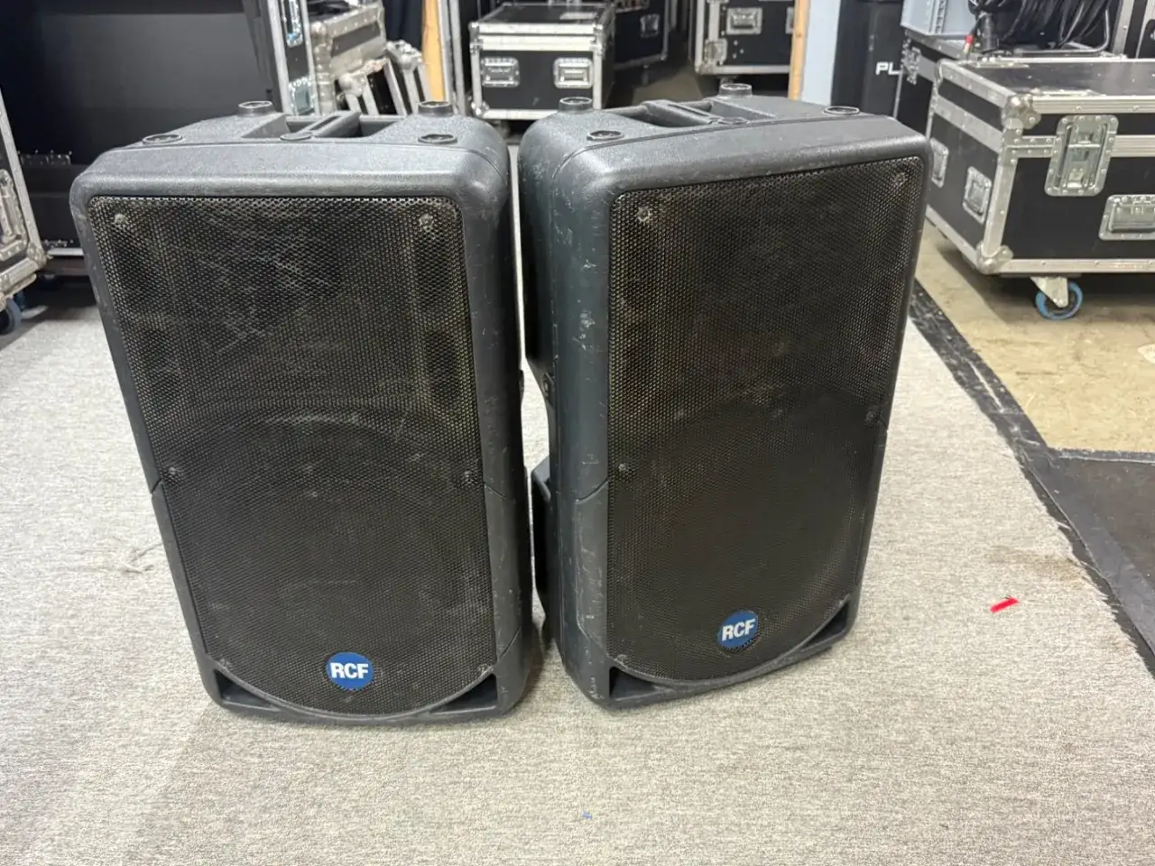 Zwei schwarze RCF 322a Lautsprecher stehen nebeneinander auf einem Teppichboden. Im Hintergrund sind weitere Flightcases zu sehen.