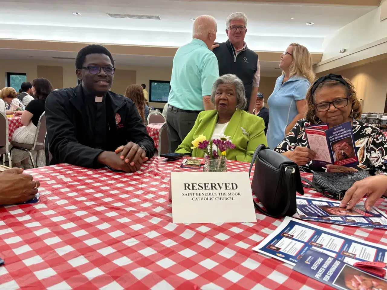 A priest, likely one of the Missionaries of Charity Fathers, smiles at a gathering. People are seated at tables covered with red and white checkered cloths.