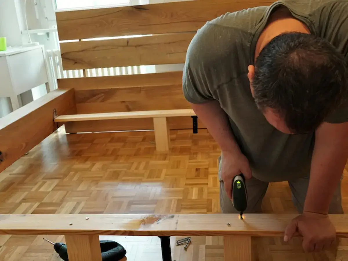 Mann schraubt am **Bett selber bauen**. Holzrahmen wird montiert, Werkzeug liegt bereit.