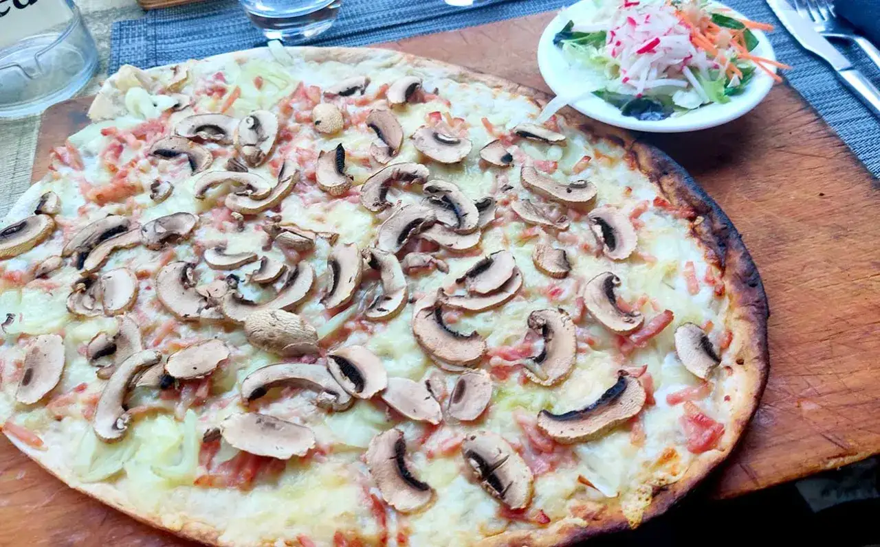 Ein köstlicher Straßburger Flammkuchen mit Pilzen und Speck, serviert auf einem Holzbrett, daneben ein kleiner Salat.