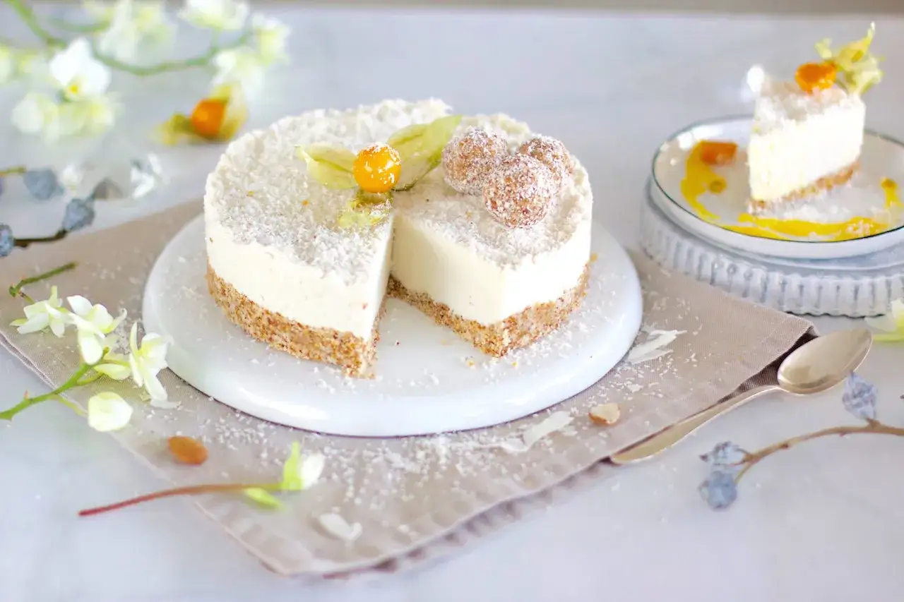 Eine Raffaello Torte, bedeckt mit Kokosraspeln und garniert mit Physalis und Kokoskugeln, steht auf einem weißen Teller.