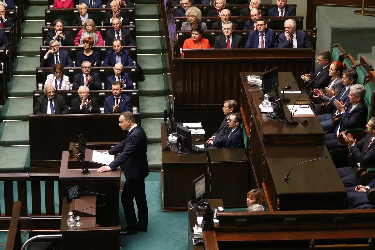 Mężczyzna przemawia z mównicy w sali sejmowej, a posłowie siedzą w ławach. To ważne orędzie.