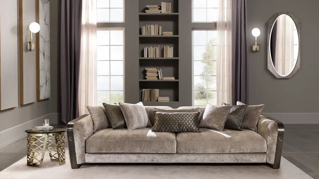 Un elegante specchio decorativo si trova dietro un divano beige, con una libreria a muro e finestre sullo sfondo.