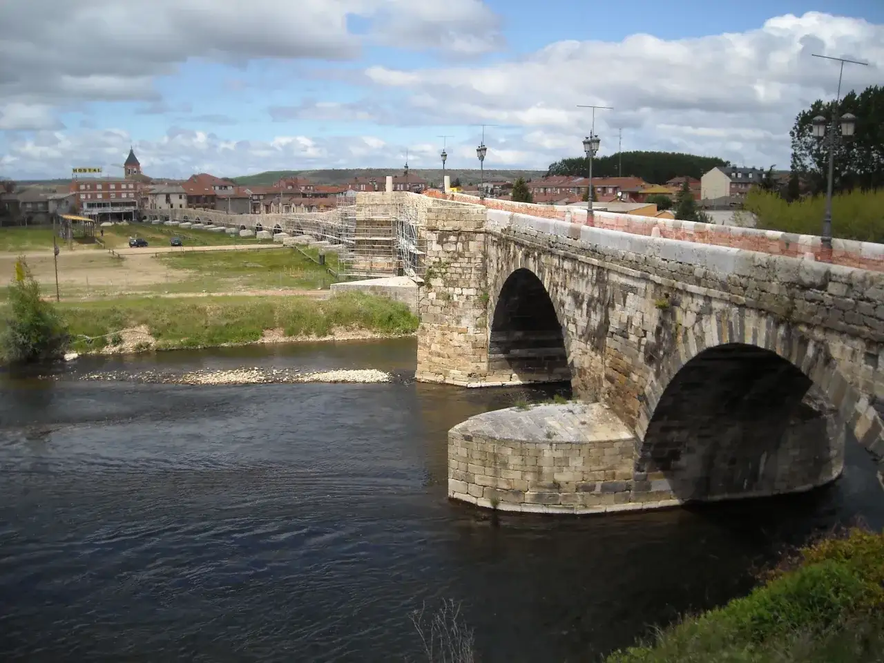Puente medieval de piedra en Hospital de Órbigo, con arcos sobre un río y edificios al fondo.