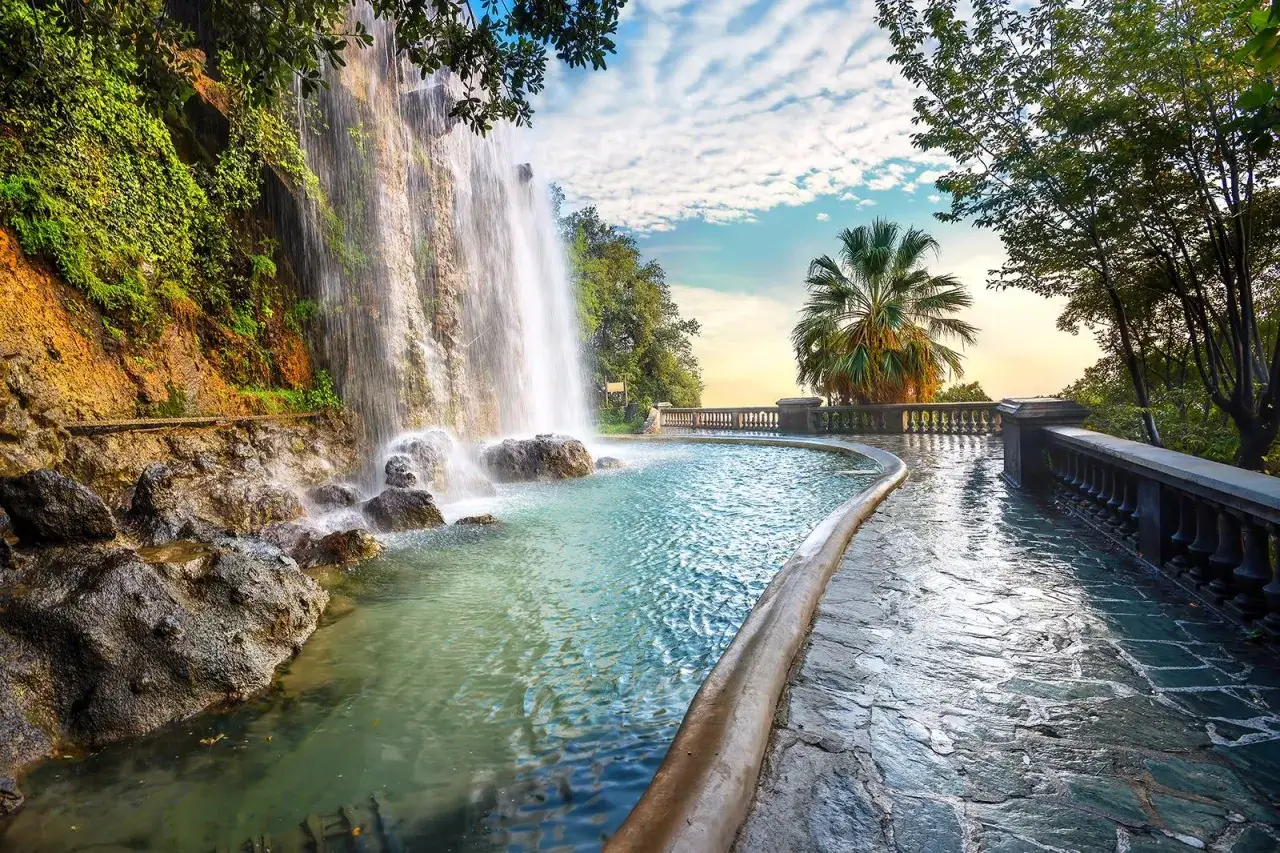 A cascading waterfall flows into a pool on Castle Hill in Nice, France, with a palm tree and cloudy sky in the background.