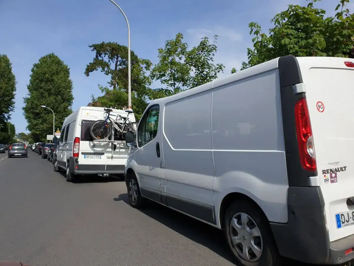 Un fourgon blanc de 6 mètres est garé dans une file de véhicules. Un autre fourgon blanc, aménagé en camping-car avec un vélo à l'arrière, est juste devant.