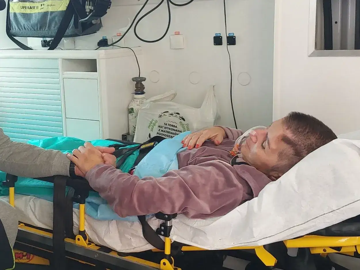 Pacjentka po pęknięciu tętniaka mózgu, w karetce, z maską tlenową, trzymająca się za rękę. Rokowania niepewne.