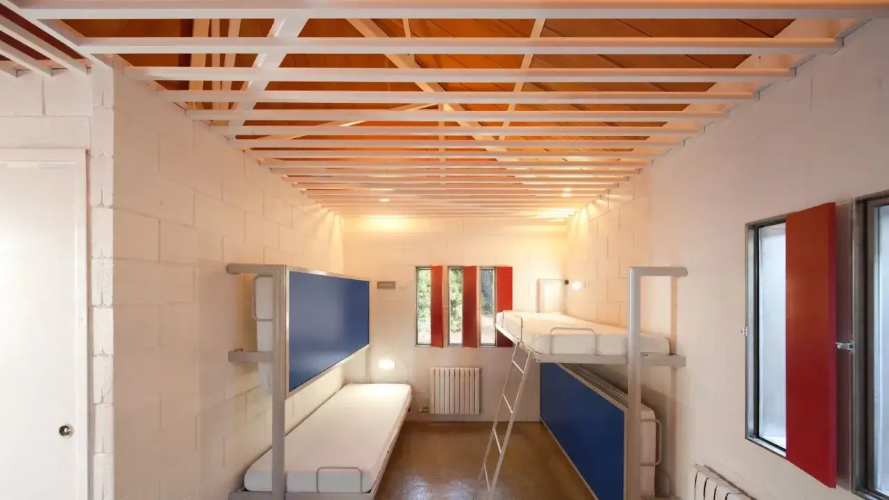 Dormitorio moderno con literas, ideal para campamentos de inglés en España. Paredes de ladrillo blanco, techo de madera y metal, ventanas con contraventanas rojas.