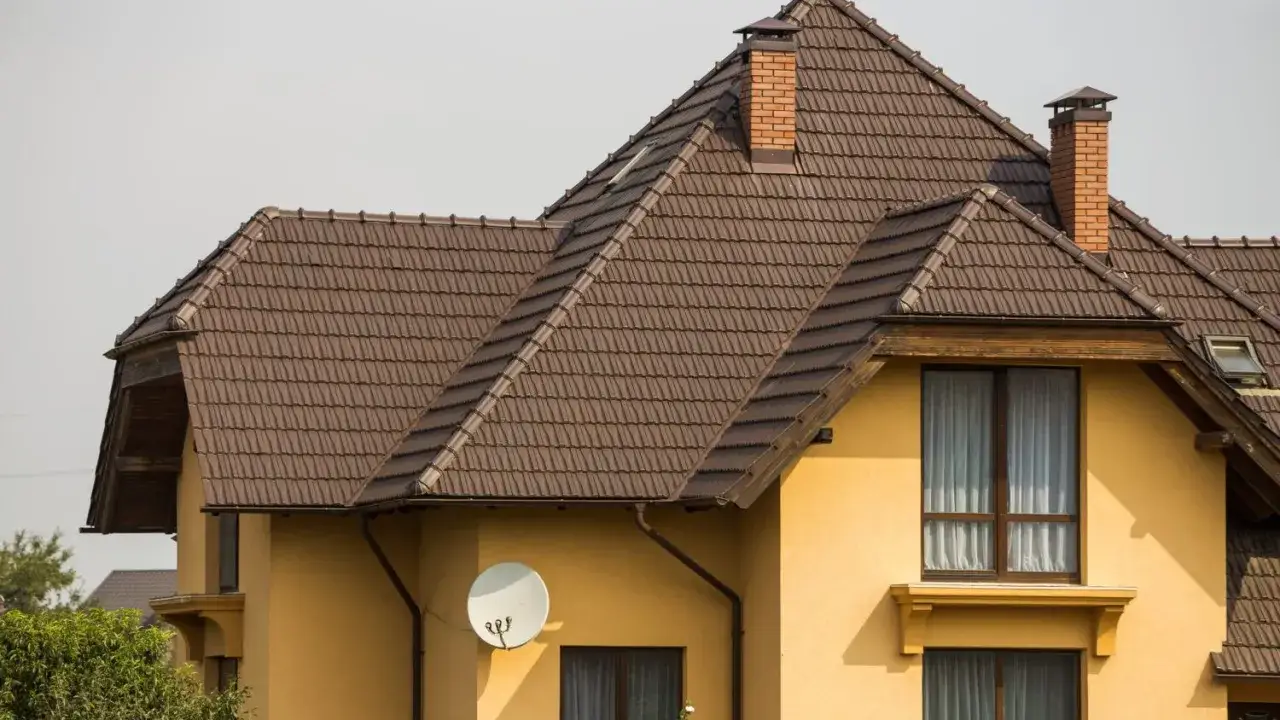 Brązowy dach nacz&oacute;łkowy z dach&oacute;wki, z ceglanym kominem i anteną satelitarną na ż&oacute;łtej ścianie domu.