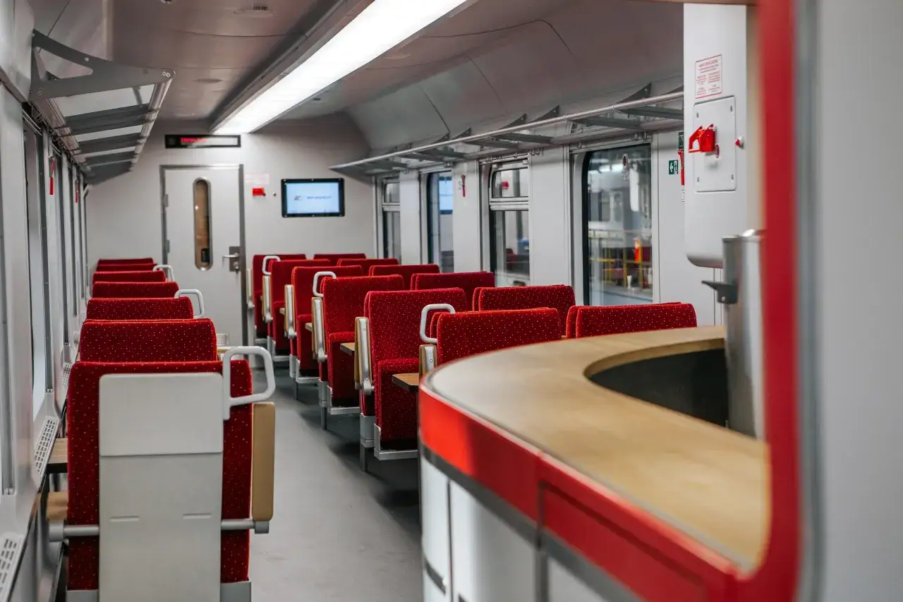 Wnętrze wagonu PKP Intercity. Puste czerwone siedzenia w rzędach, stoliki i ekran informacyjny.