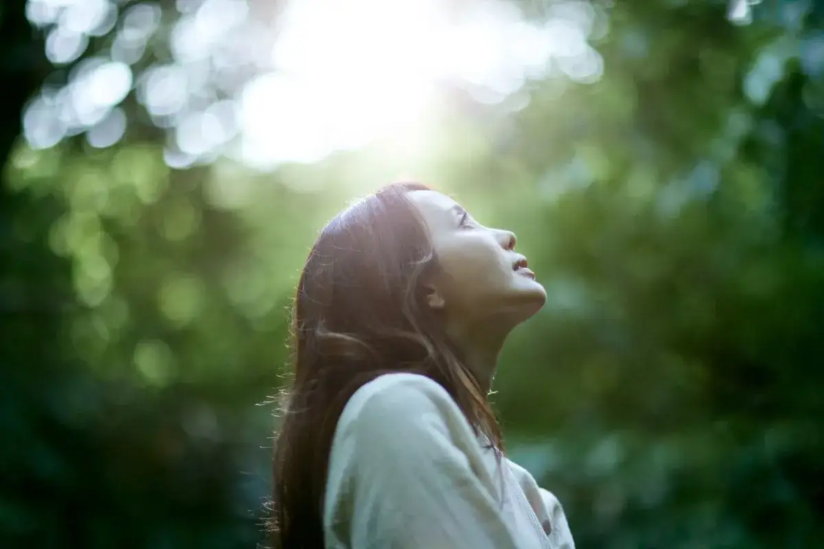 A woman looks up at the sunlit sky, embracing the art of doing nothing amidst a soft, green, bokeh background.