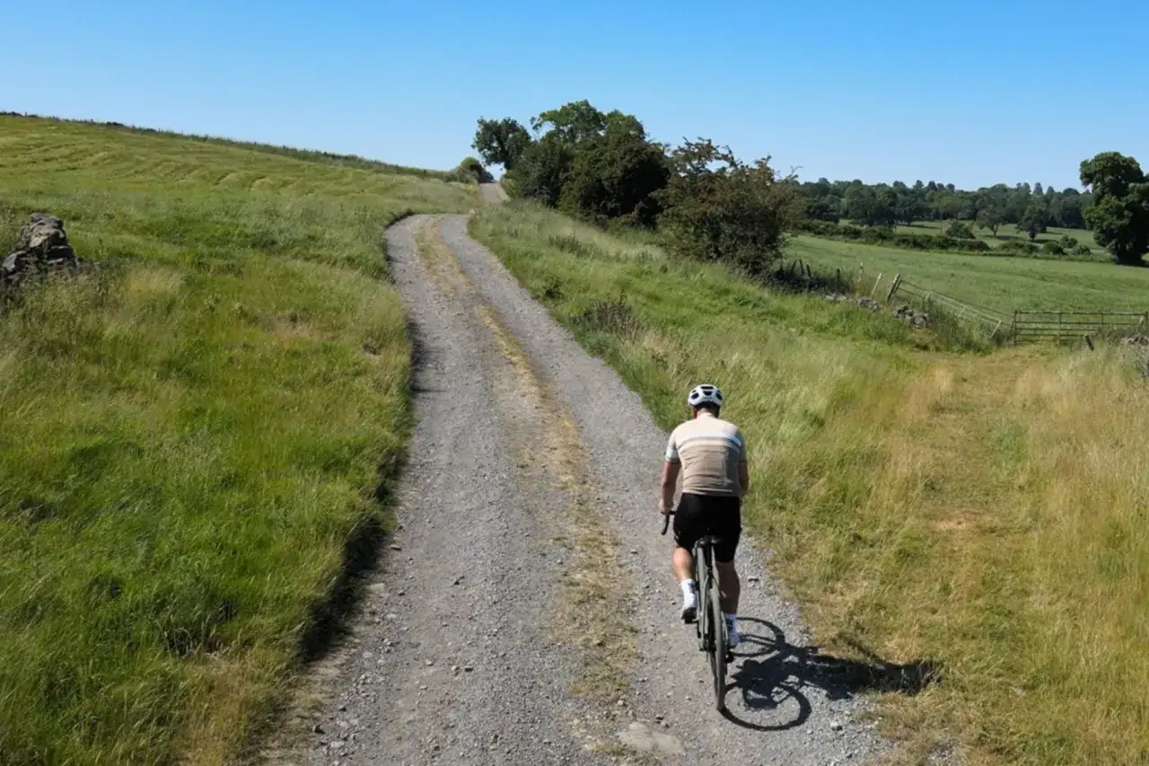 A cyclist rides up a gravel path, followed by a drone, through rolling green hills under a clear blue sky.