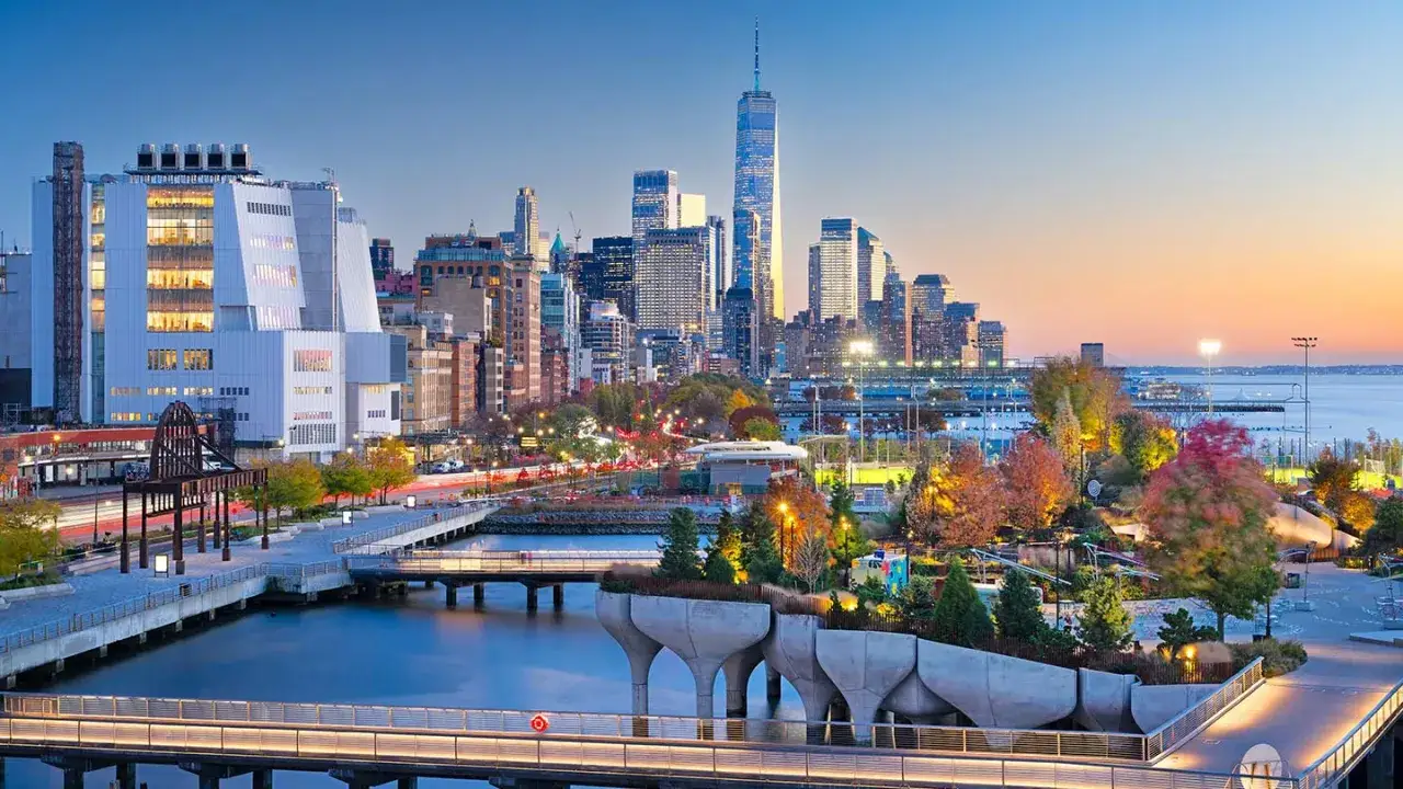 Solnedgång över Big Apple New York. En modern park med träd och gångvägar vid vattnet, med skyskrapor i bakgrunden.