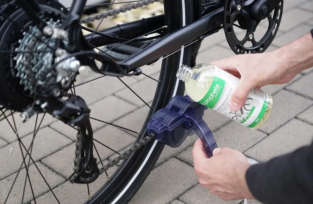 Fahrradkette reinigen mit speziellem Entfetter. Ein Werkzeug reinigt die Kette, w&auml;hrend eine Hand die Fl&uuml;ssigkeit aus einer Flasche aufgie&szlig;t.