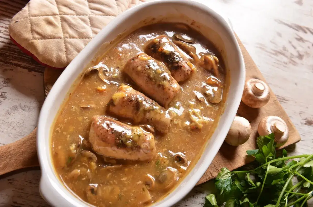 Pyszny schab z pieczarkami w sosie, podany w białej owalnej misie. Obok świeże pieczarki i natka pietruszki.