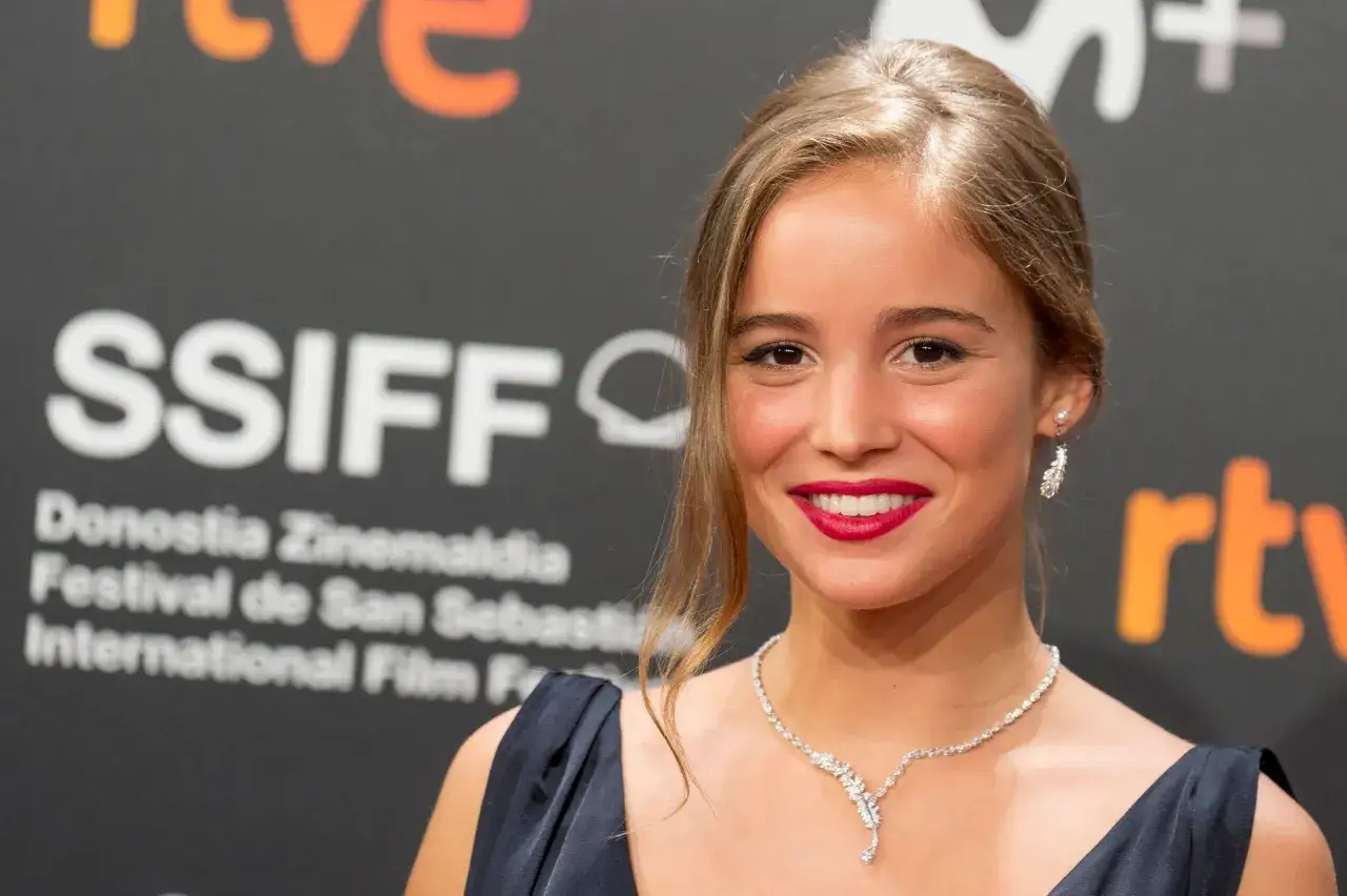 Alba Baptista ler vackert p&aring; r&ouml;da mattan, kl&auml;dd i en elegant kl&auml;nning och glittrande smycken.