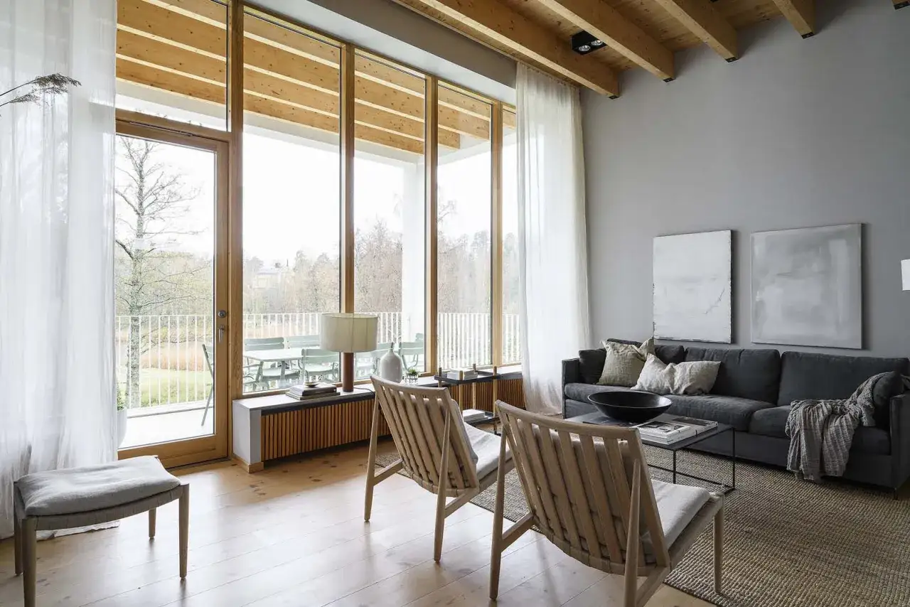 Gem&uuml;tliches Wohnzimmer mit warmes grau farbpalette, Holzbalken und gro&szlig;en Fenstern, die auf eine Terrasse mit Blick ins Gr&uuml;ne f&uuml;hren.