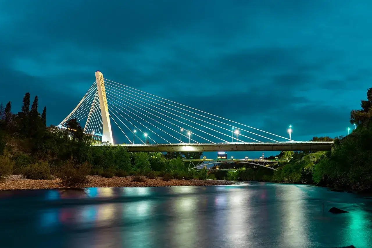 Podgorica co zobaczyć: nowoczesny most nad rzeką o zmierzchu, z odbiciami świateł na wodzie.