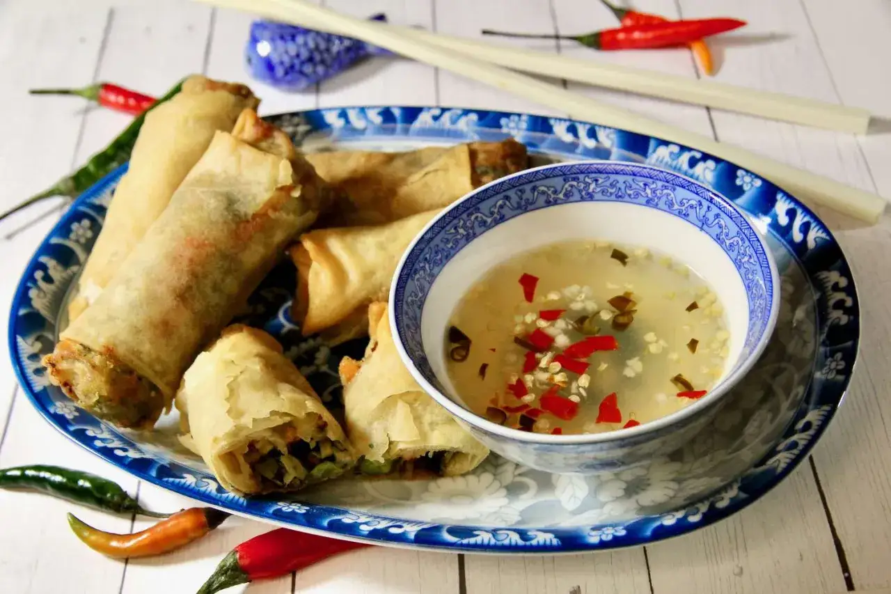 Chrupiące sajgonki na talerzu z niebieskim wzorem, obok miseczka z pikantnym sosem do sajgonek, ozdobiona papryczkami chili.