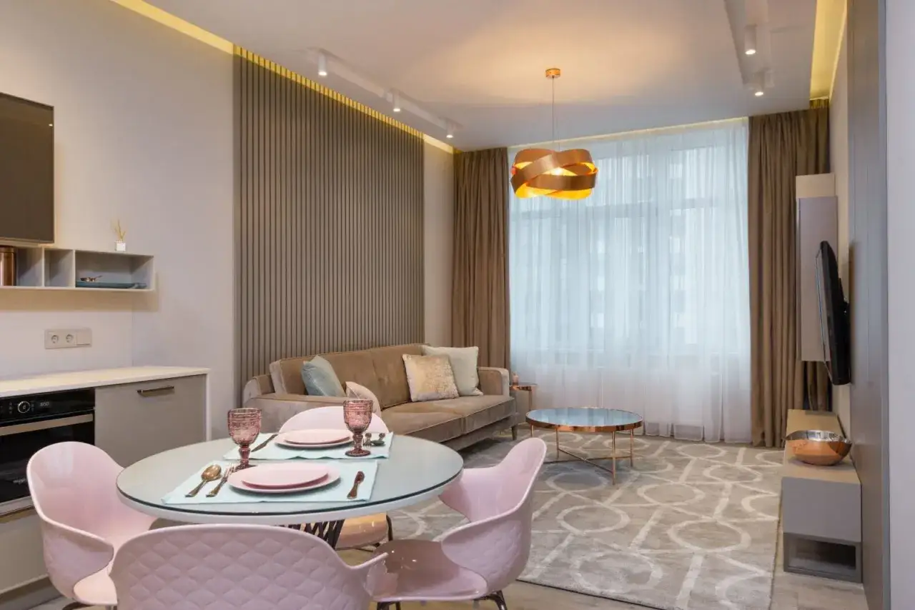 Nowoczesny salon z aneksem kuchennym po generalnym remoncie mieszkania 50m2. Jasne kolory, welurowa sofa, okrągły stół z różowymi krzesłami.