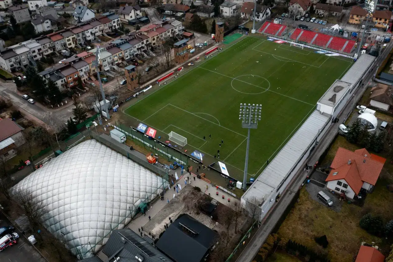 Widok z lotu ptaka na niepołomice stadion z balonem pneumatycznym obok. Boisko jest zielone, z zaznaczonymi liniami i bramkami.