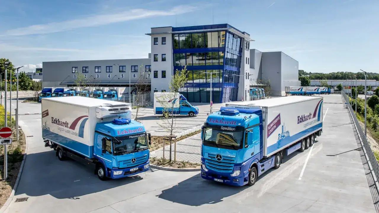 Zwei blaue LKW der Spedition Eckhardt stehen vor einem modernen Logistikzentrum. Das Bild zeigt, was eine Spedition leistet: Transport und Logistik.