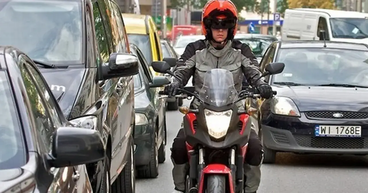 Motocyklista w kasku i kurtce próbuje przejechać między samochodami w korku. Czy motor może jechać między samochodami? Tak, jeśli jest to bezpieczne.