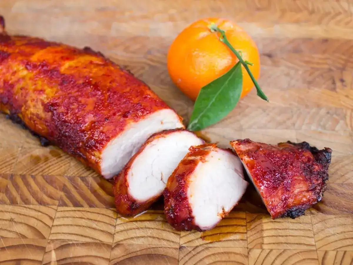 Un filet de porc fum&eacute;, laqu&eacute; et tranch&eacute;, repose sur une planche &agrave; d&eacute;couper en bois, &agrave; c&ocirc;t&eacute; d'une mandarine fra&icirc;che.