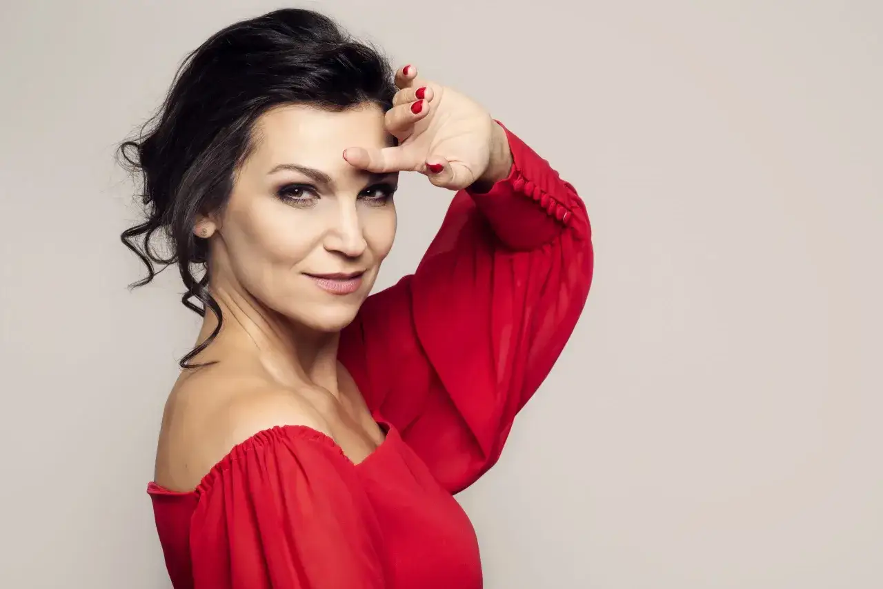 Olga Bończyk w czerwonej sukni, z dłonią przy czole, jakby wypatrywała swojego męża.