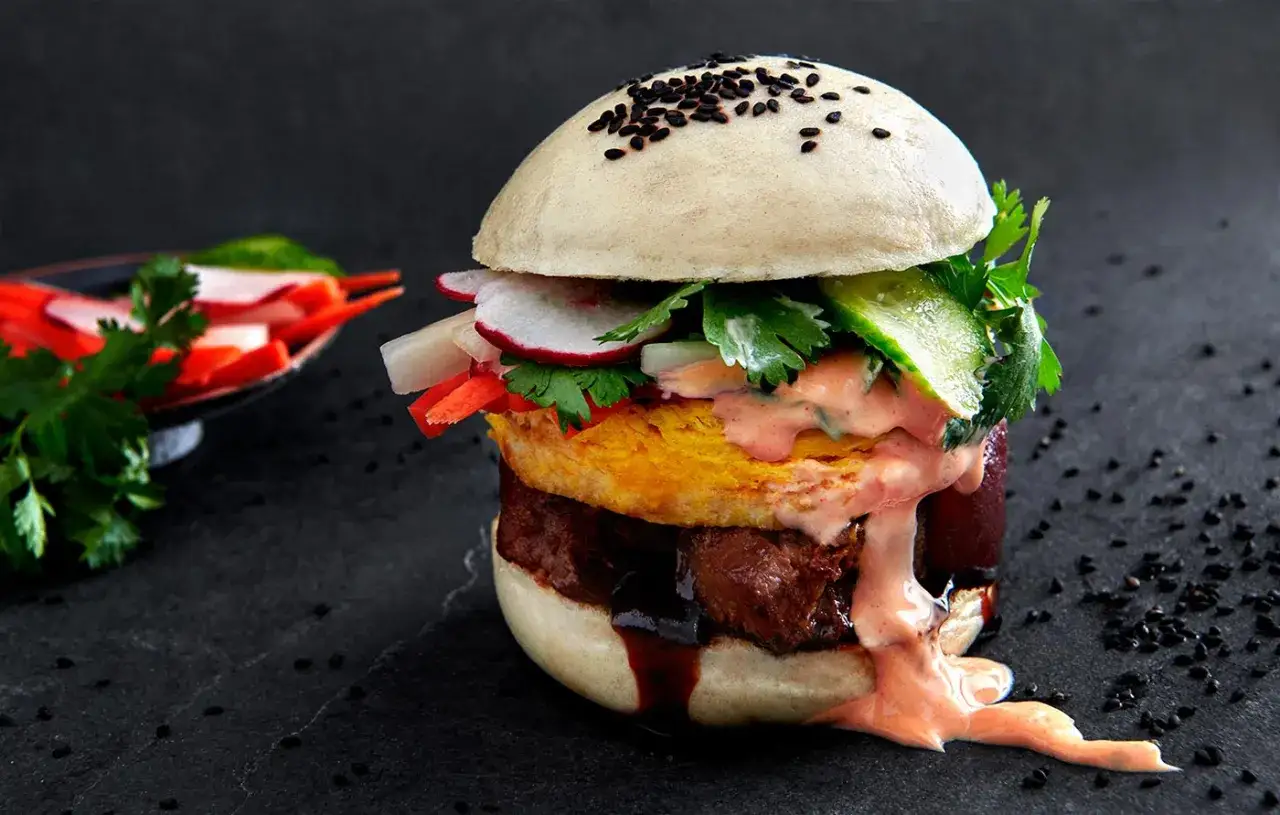 Ein saftiger bao burger mit Fleisch, Ei, Gurke, Radieschen und einer cremigen Sauce, bestreut mit schwarzen Sesamk&ouml;rnern.
