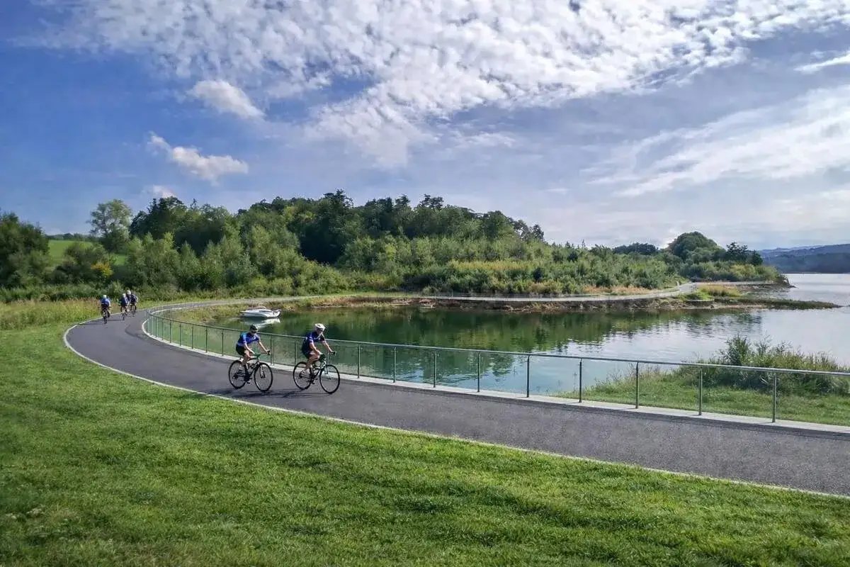 Grupa rowerzystów przemierza malowniczą trasę nad jeziorem. W tle zielone wzgórza i błękitne niebo. Idealne miejsce na **velo skawa** i aktywny wypoczynek.