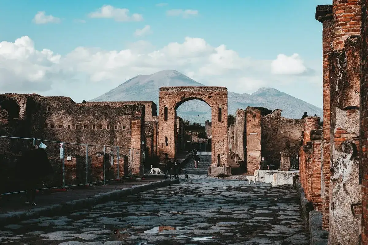 Ancient Italian ruins stand under a cloudy sky, with Mount Vesuvius looming in the distance. Cobblestone streets reflect the light.