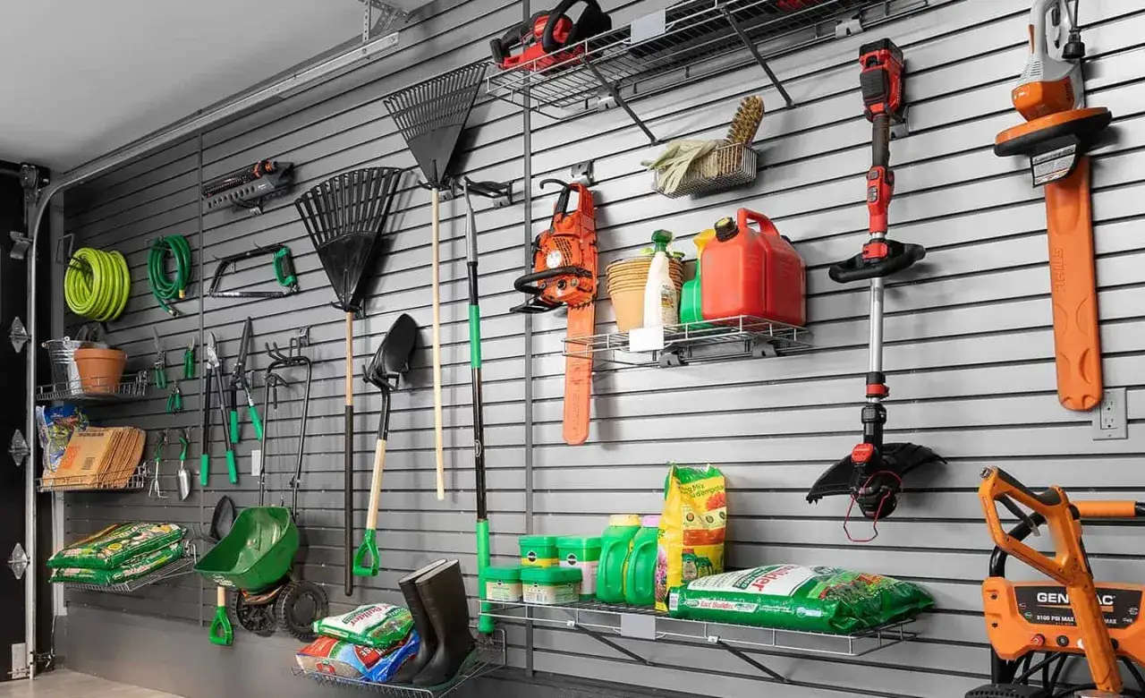 Un mur de garage organis&eacute; avec une astuce rangement outils garage : r&acirc;teaux, tron&ccedil;onneuse, d&eacute;broussailleuse, tuyau d'arrosage, pelles, et sacs d'engrais.