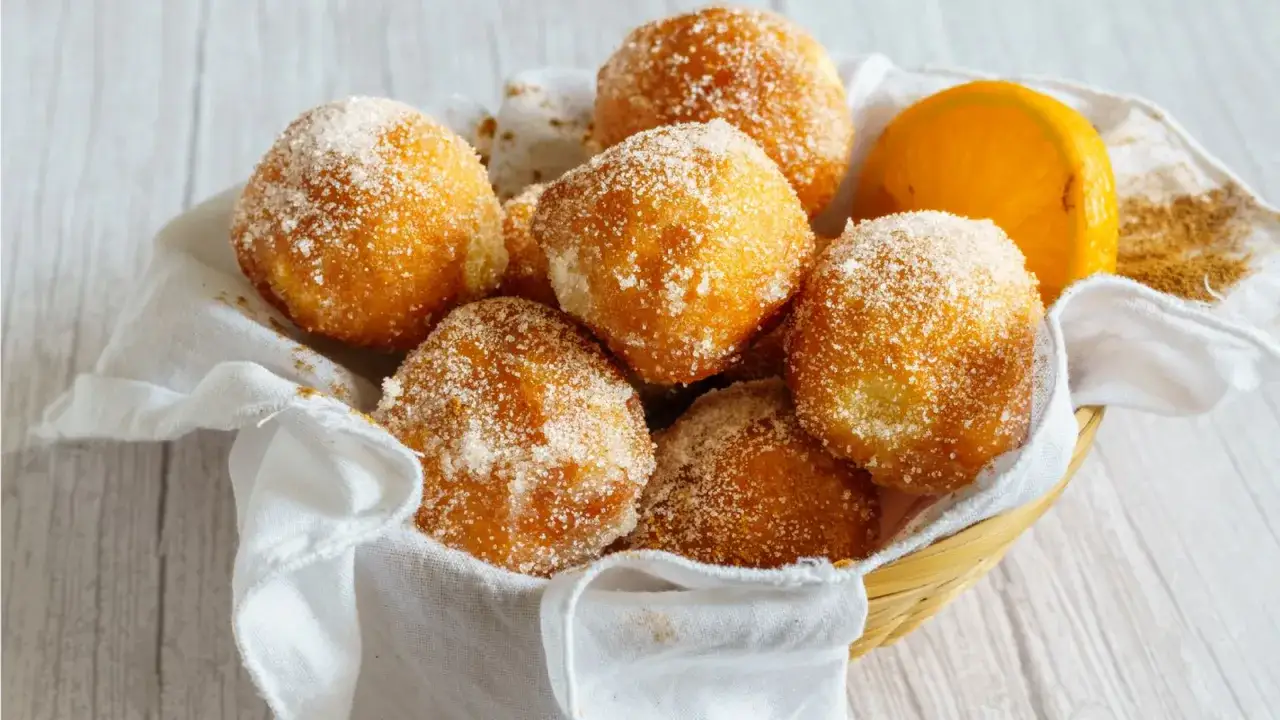 Cesta de mimbre con buñuelos de viento espolvoreados con azúcar y canela, junto a una rodaja de naranja.