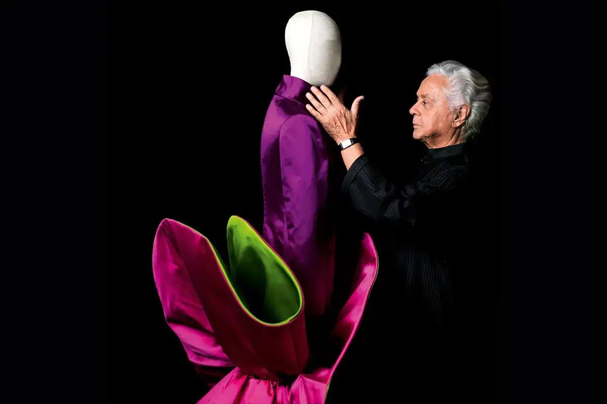 Roberto Capucci, maestro della moda, accarezza un abito viola su un manichino, con un'esplosione di petali fucsia e verdi ai suoi piedi.