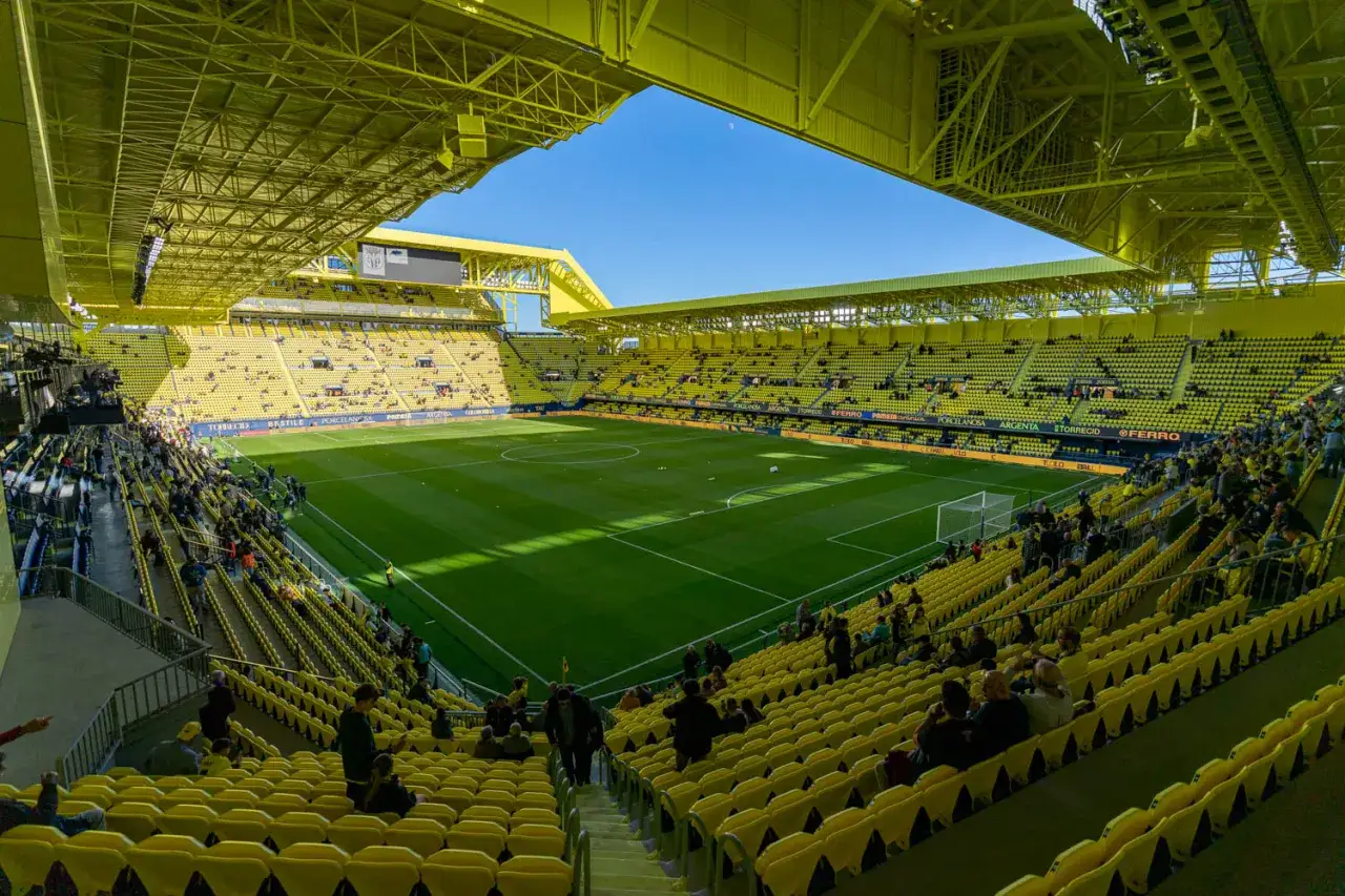 Stadion Villarreal, pełen żółtych krzesełek, z zieloną murawą i błękitnym niebem.