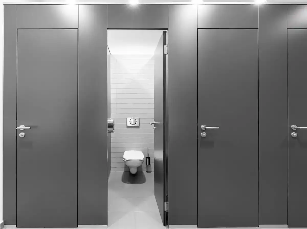 Reihe von Toilettenkabinen mit grauen Türen. Eine Tür ist geöffnet und gibt den Blick auf eine Toilette frei. Die Frage