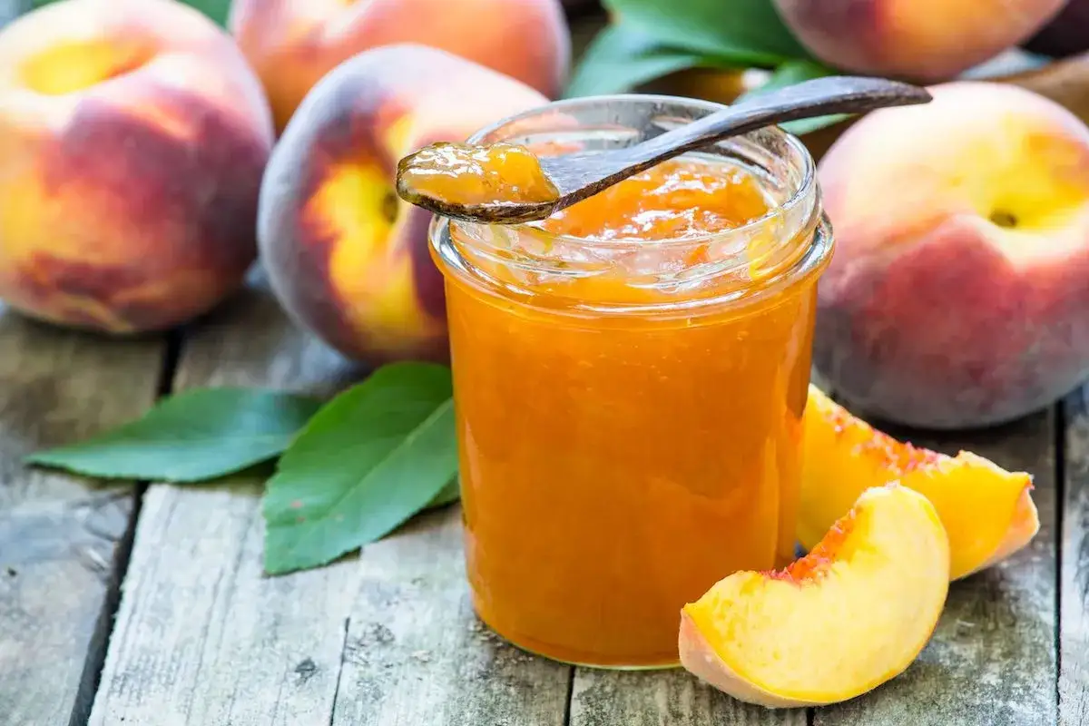 Barattolo di marmellata di pesche su un tavolo di legno, con pesche fresche e foglie verdi intorno.