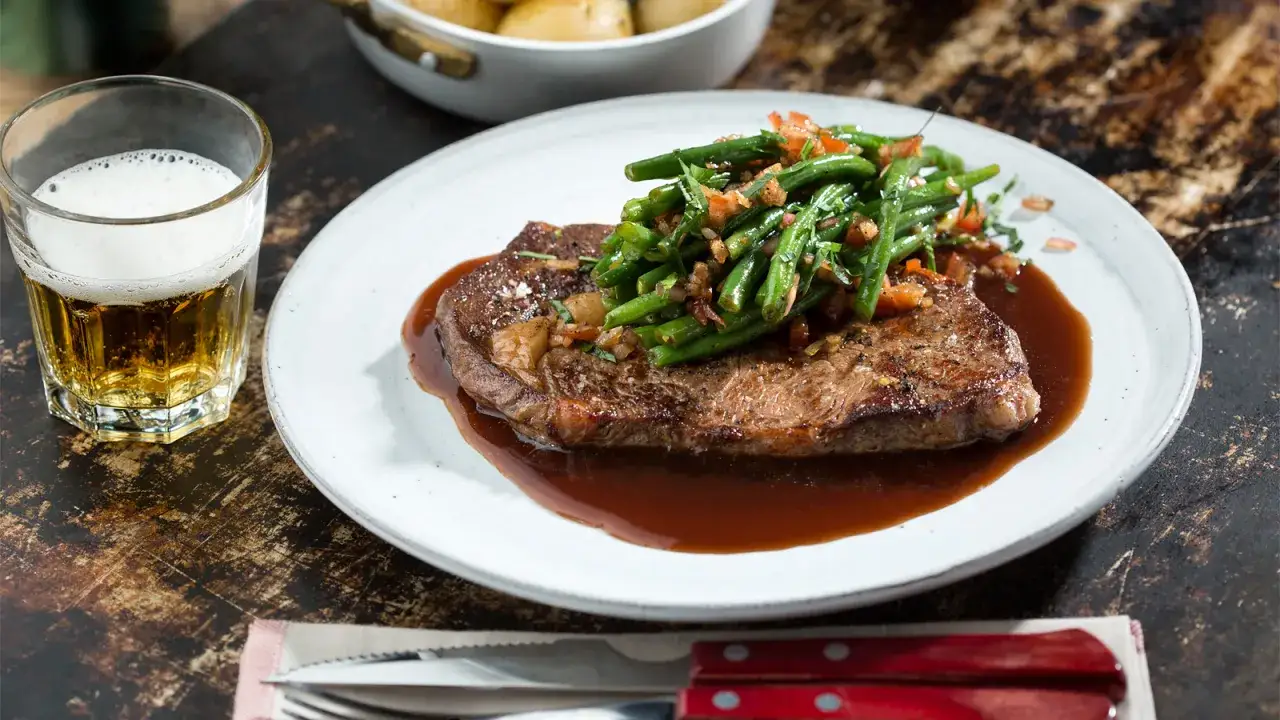 En saftig entrecote med gröna bönor och en mustig sås, perfekt för ett glas vin till entrecote. En öl står bredvid.