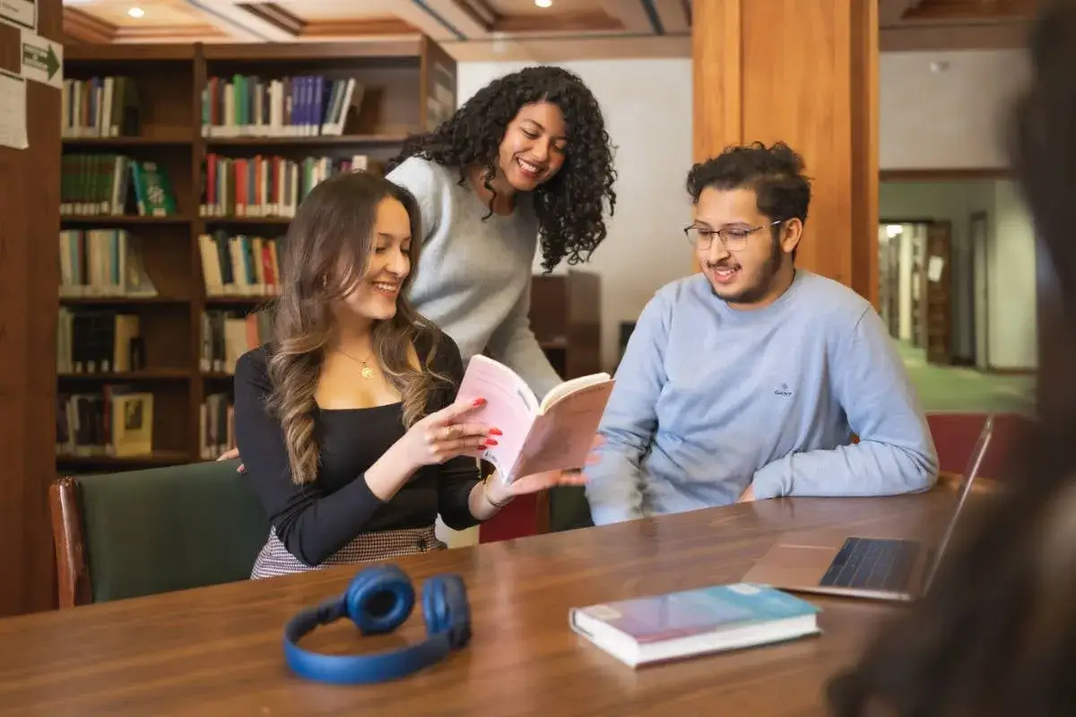 Trois &eacute;tudiants, dont un master moyen orient, &eacute;tudient ensemble dans une biblioth&egrave;que, partageant un livre et un ordinateur portable.