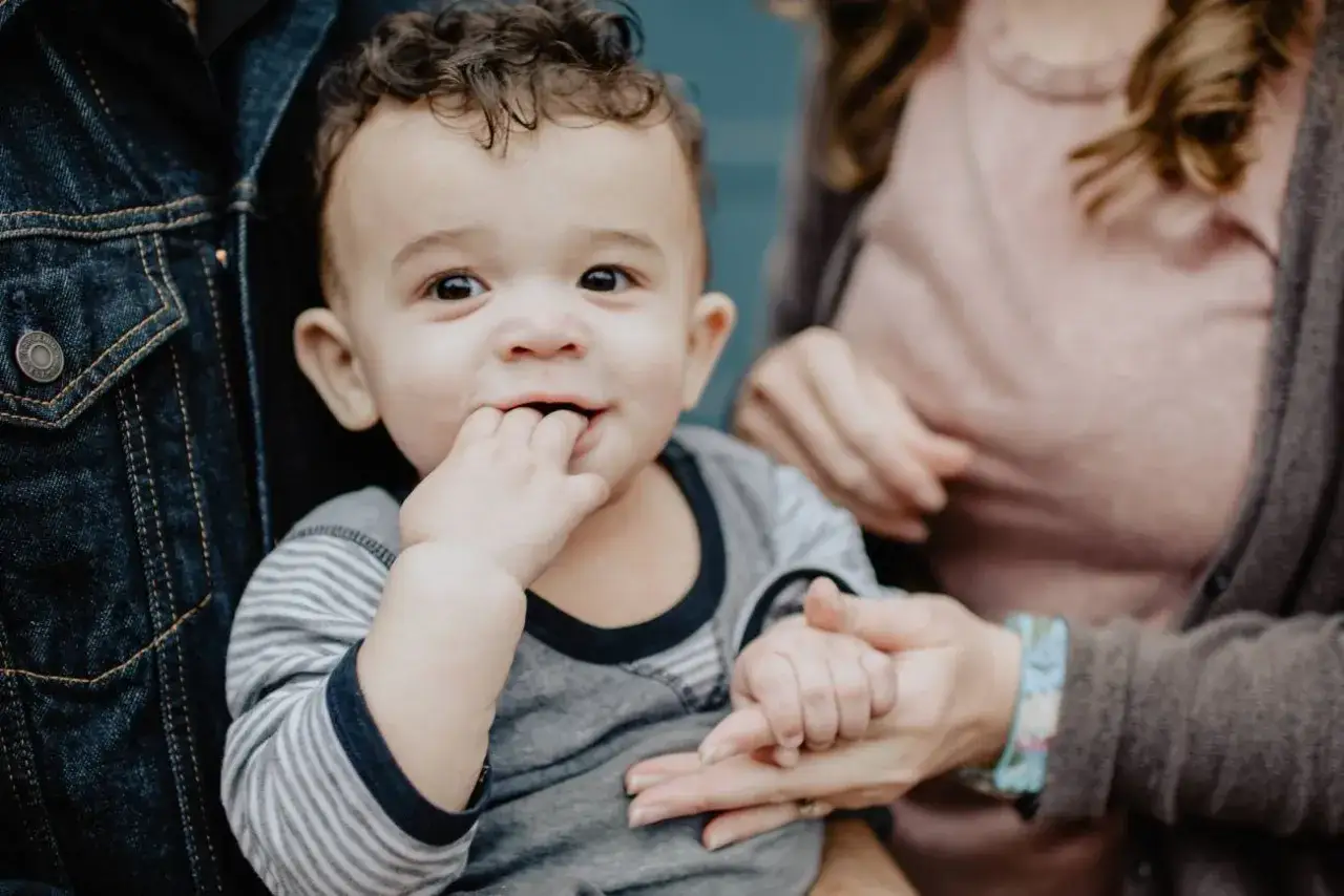 Uśmiechnięte dziecko z kręconymi włosami wkłada paluszki do buzi, trzymane przez rodziców. To ten magiczny moment, kiedy dziecko świadomie mówi mama.